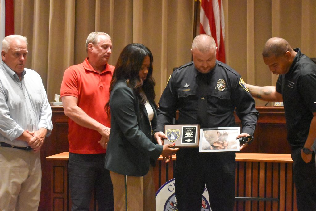 Five people stand together during a ceremony in a formal room. A police officer in uniform holds a plaque and a photo of a German Shepherd dog, while a woman next to him helps support the plaque. The three other men stand solemnly nearby, one of them placing a hand on the officer’s shoulder.