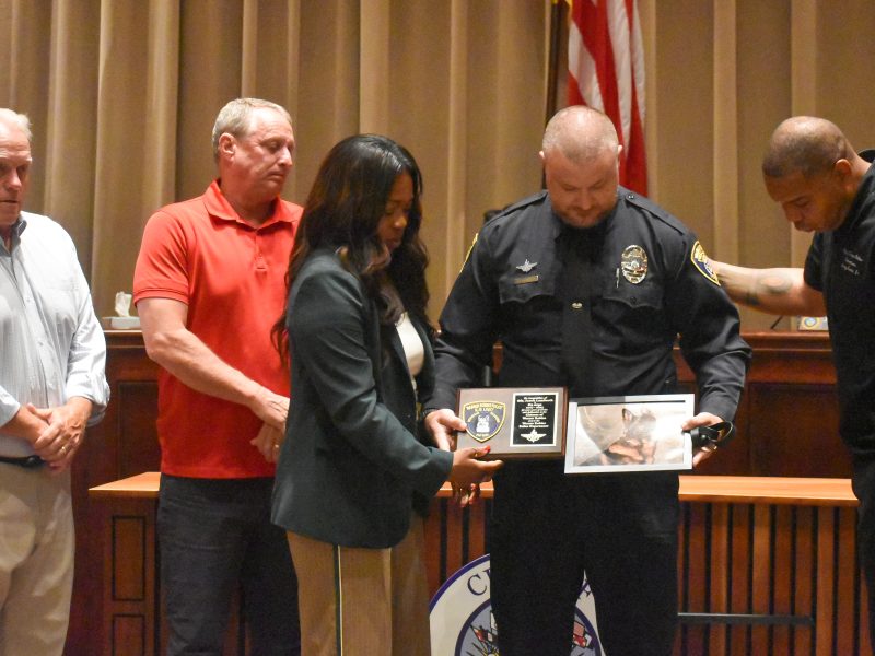 Five people stand together during a ceremony in a formal room. A police officer in uniform holds a plaque and a photo of a German Shepherd dog, while a woman next to him helps support the plaque. The three other men stand solemnly nearby, one of them placing a hand on the officer’s shoulder.