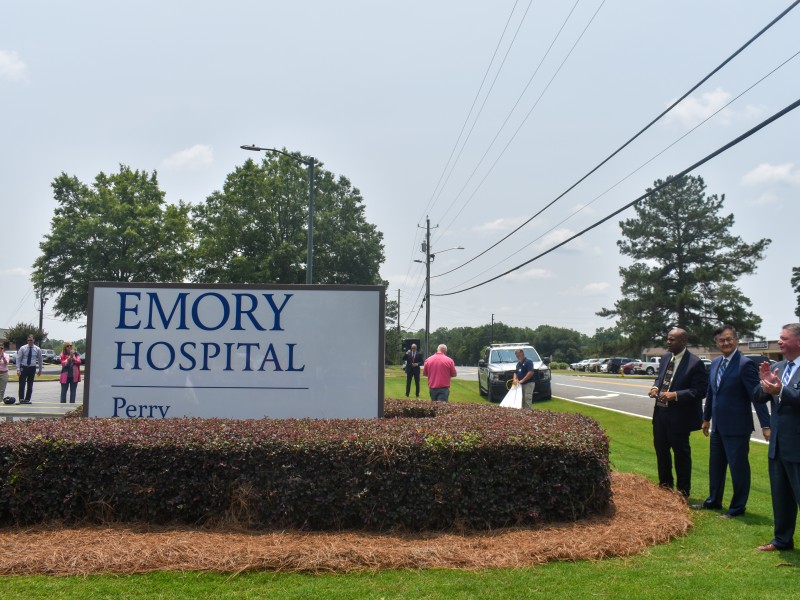 A white sign with blue letters saying "Emory Hospital." Three men are standing beside the sign smiling and clapping.