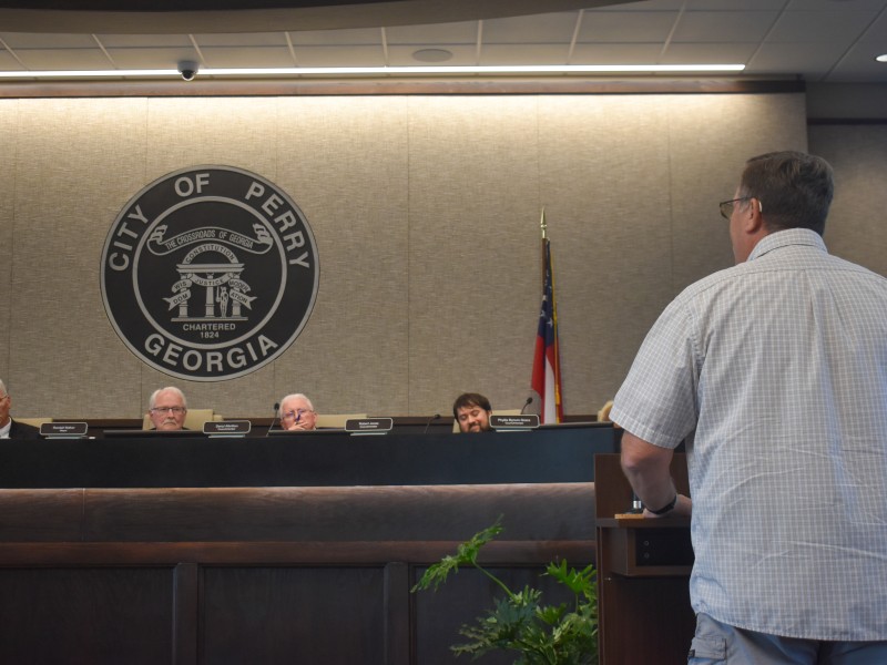 A man in a plaid shirt standing. Back is facing the camera. The man is speaking to four men who are sitting behind a table.