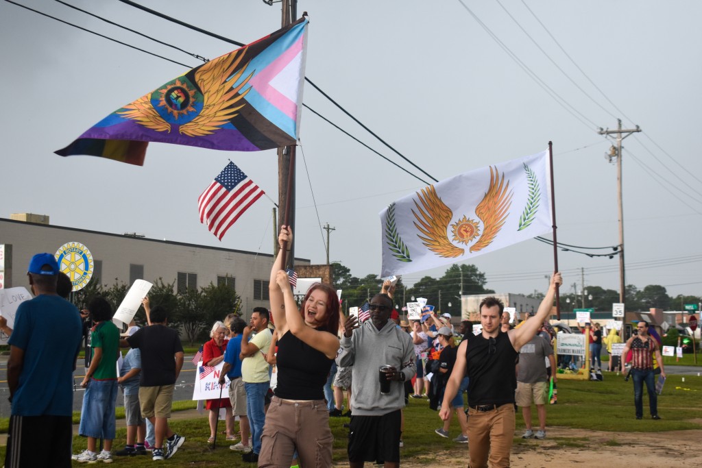 A woman and a man holding up flags. One of the flags symbolizes the LGBTQ+ and the other symbolizes solidarity. Both flags have a fist in the middle. In the background, an American flag is seen.