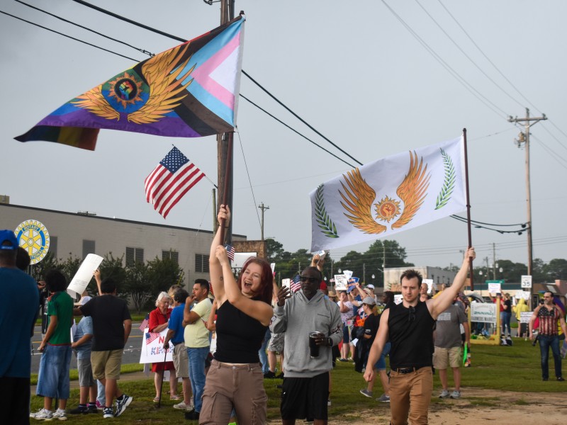 A woman and a man holding up flags. One of the flags symbolizes the LGBTQ+ and the other symbolizes solidarity. Both flags have a fist in the middle. In the background, an American flag is seen.