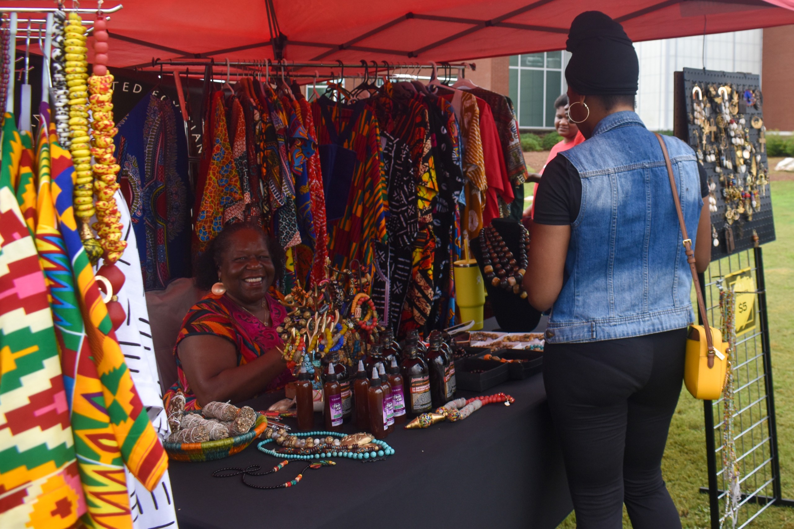 A woman smiling and sitting down. Behind her are colorful clothing hanged. In from of her is a woman looking at items on the table in front of her.