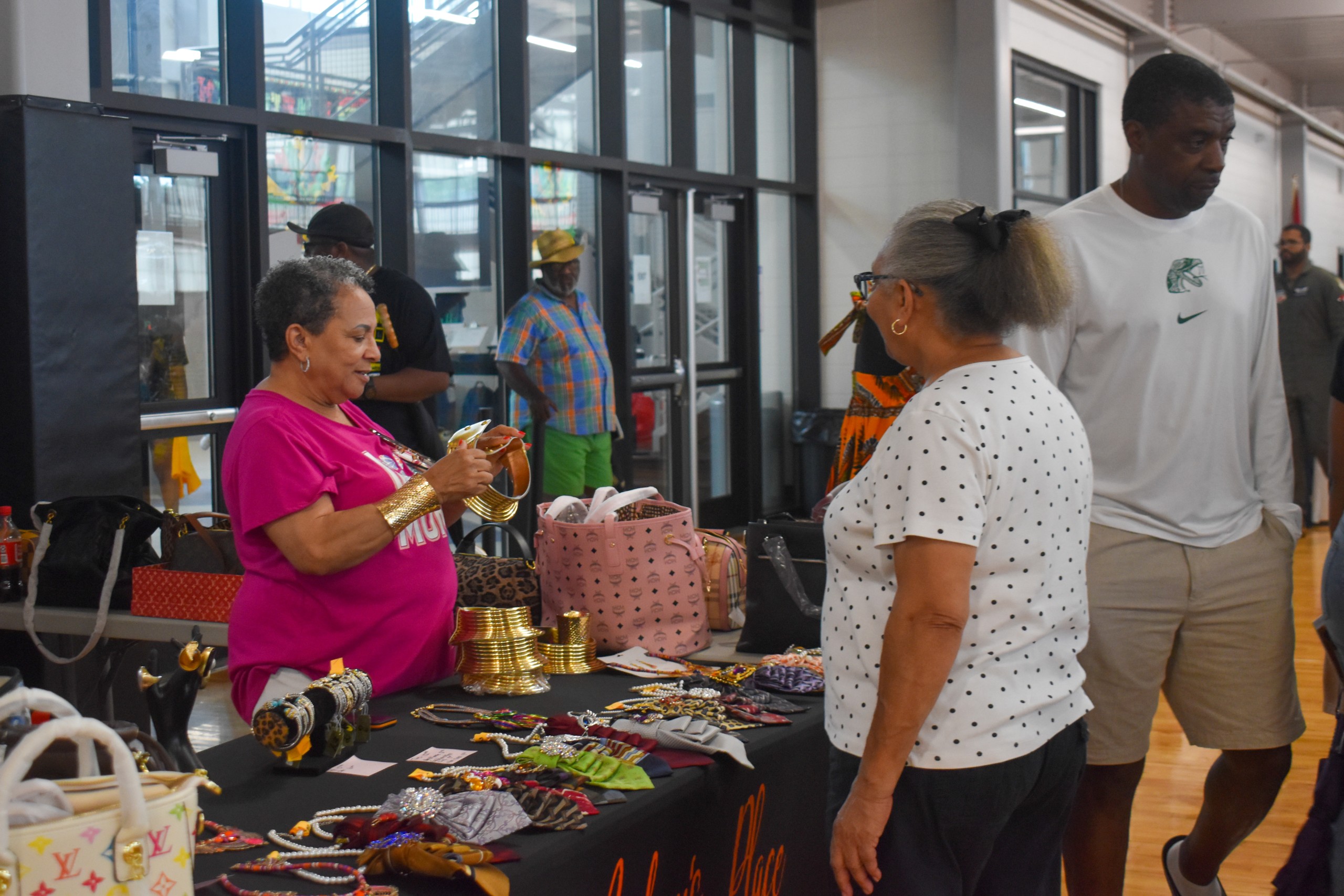 A woman in a pink shirt selling items behind a table. She is talking to a customer, a woman in a white shirt standing in front of her.