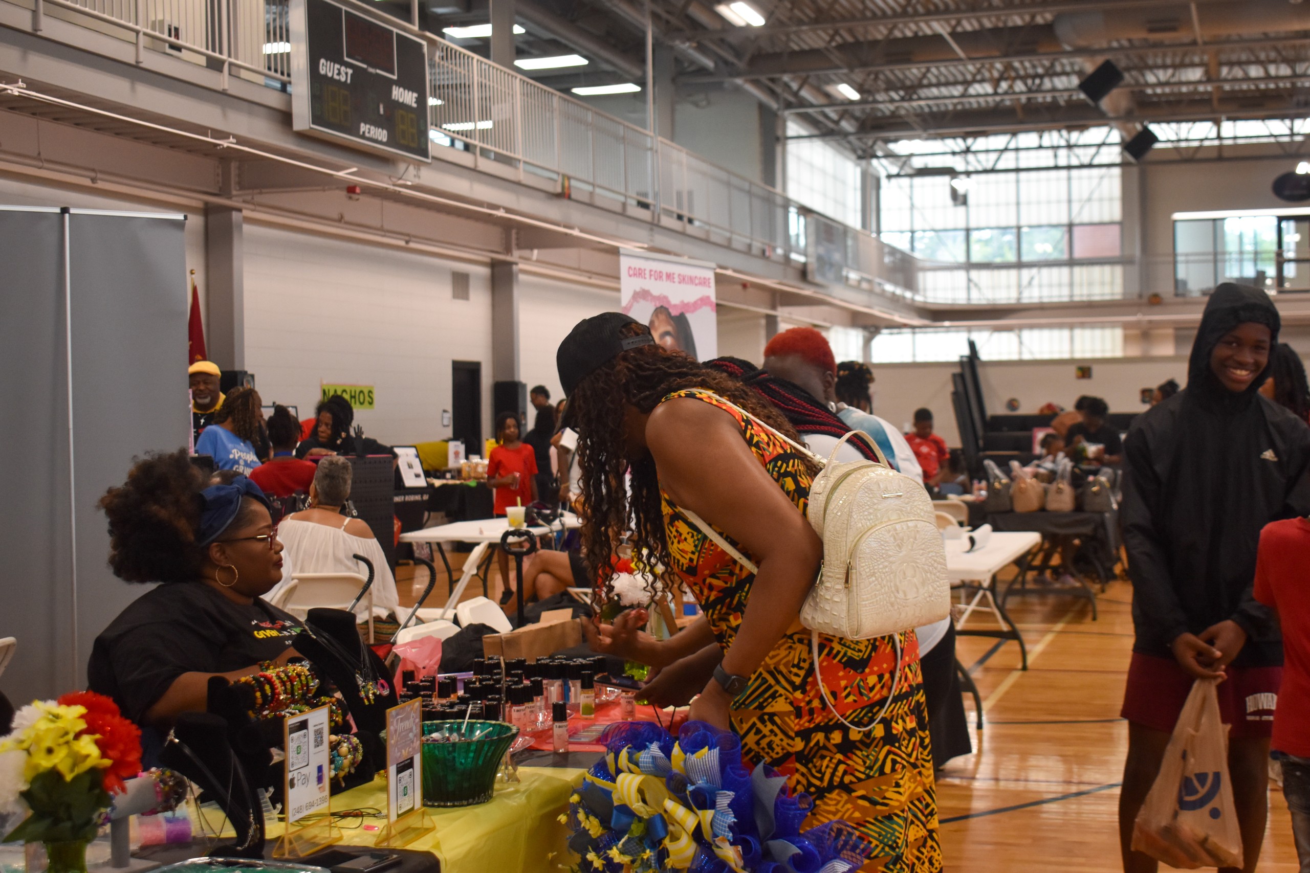 A woman in a colorful dress looking at items on a table. A boy is smiling in the background.
