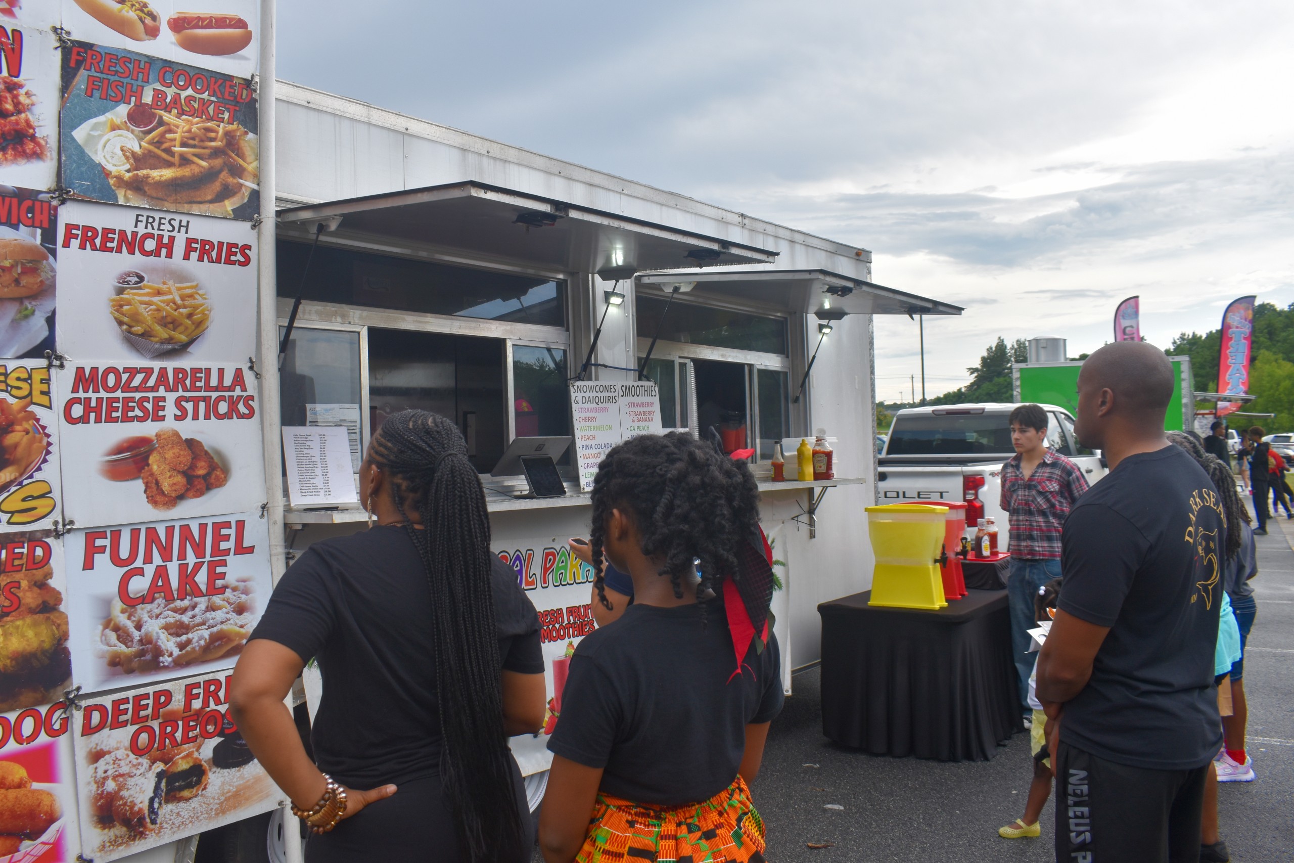 People lining up in front of a white food truck. People are reading signs of what the food truck sells. The signs have pictures of food and red letters of what the food is.