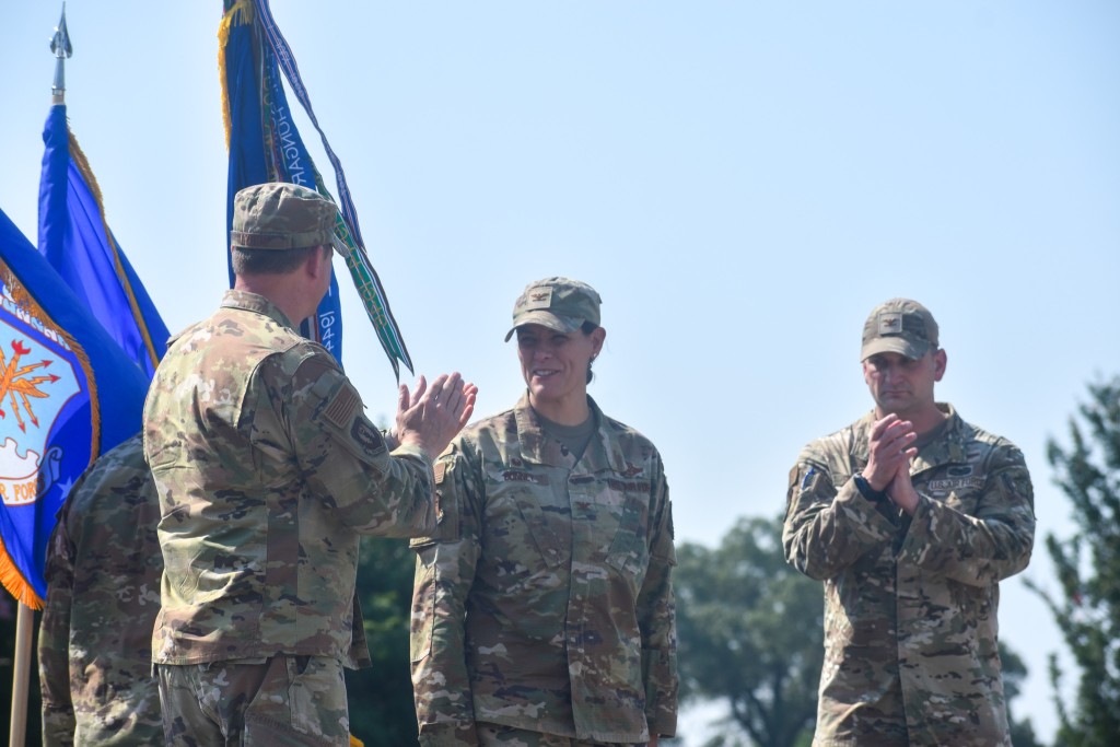 Three people in military uniform. A woman is in the middle smiling. A man has a back towards the camera and is clapping. A man is behind the woman also clapping.