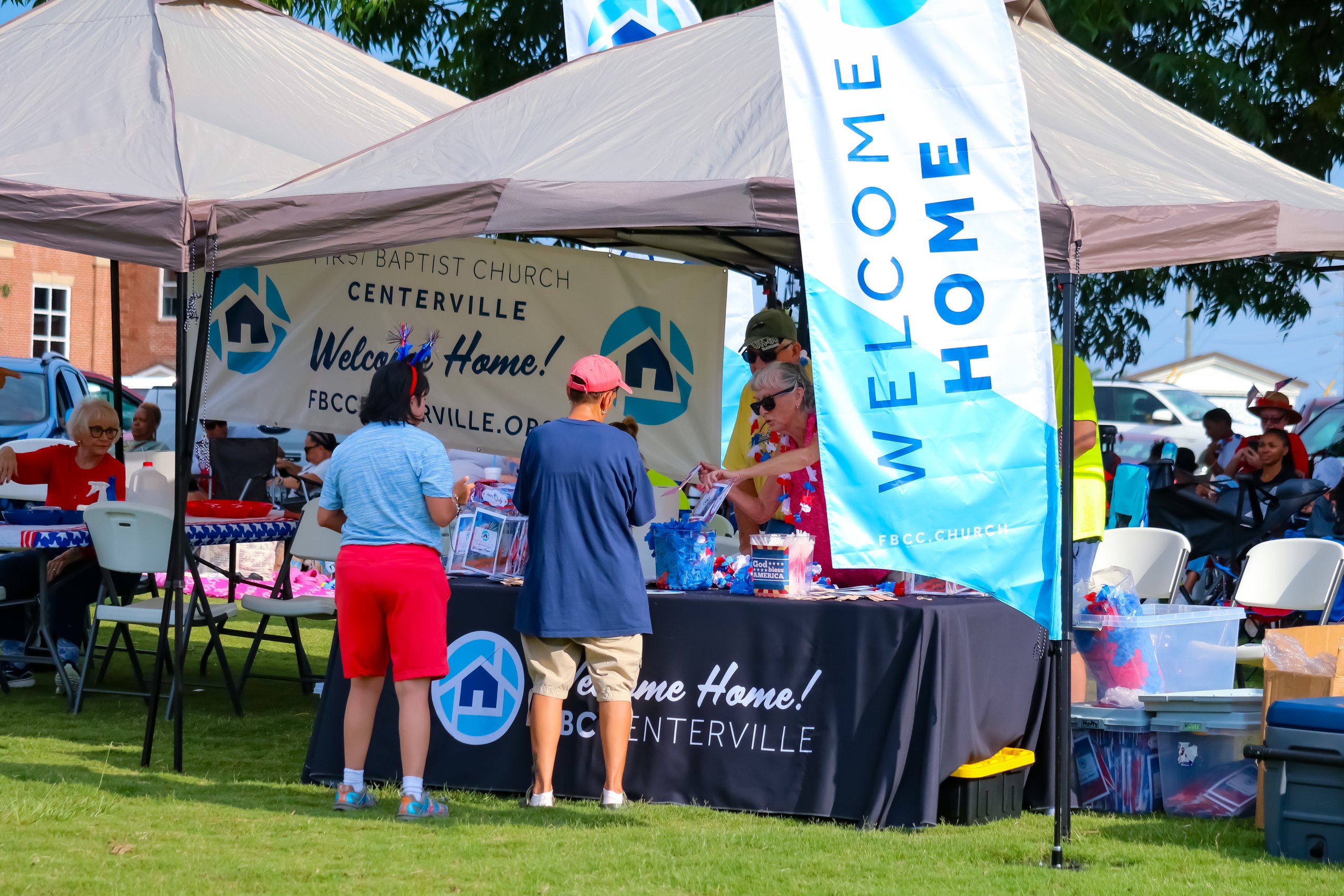 A Centerville welcome table with blue signs. On the sign, it is written "Welcome Home." People are standing at the table.
