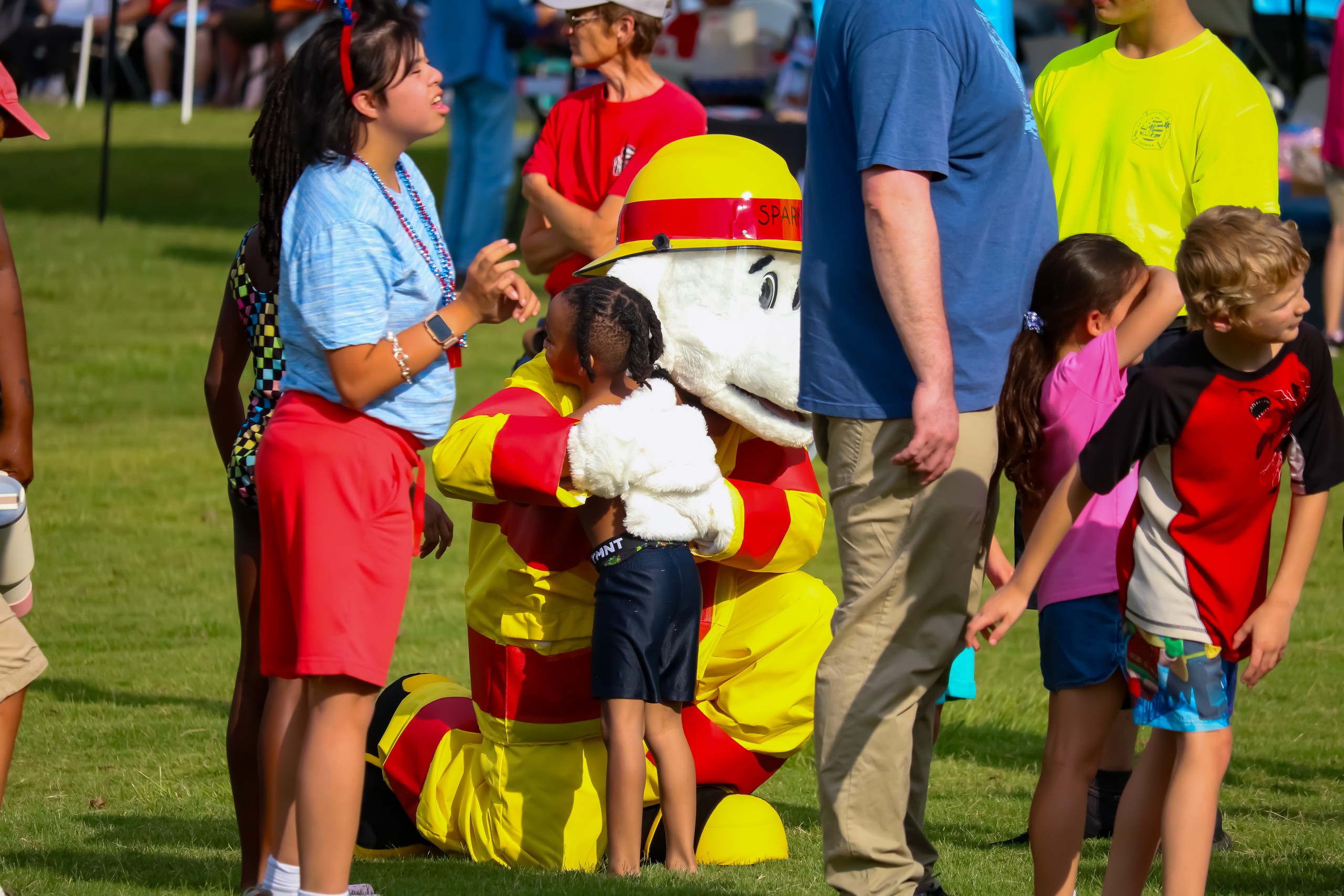 A child hugging a person in a costume. The costume is of a white dog wearing a firefighter uniform.