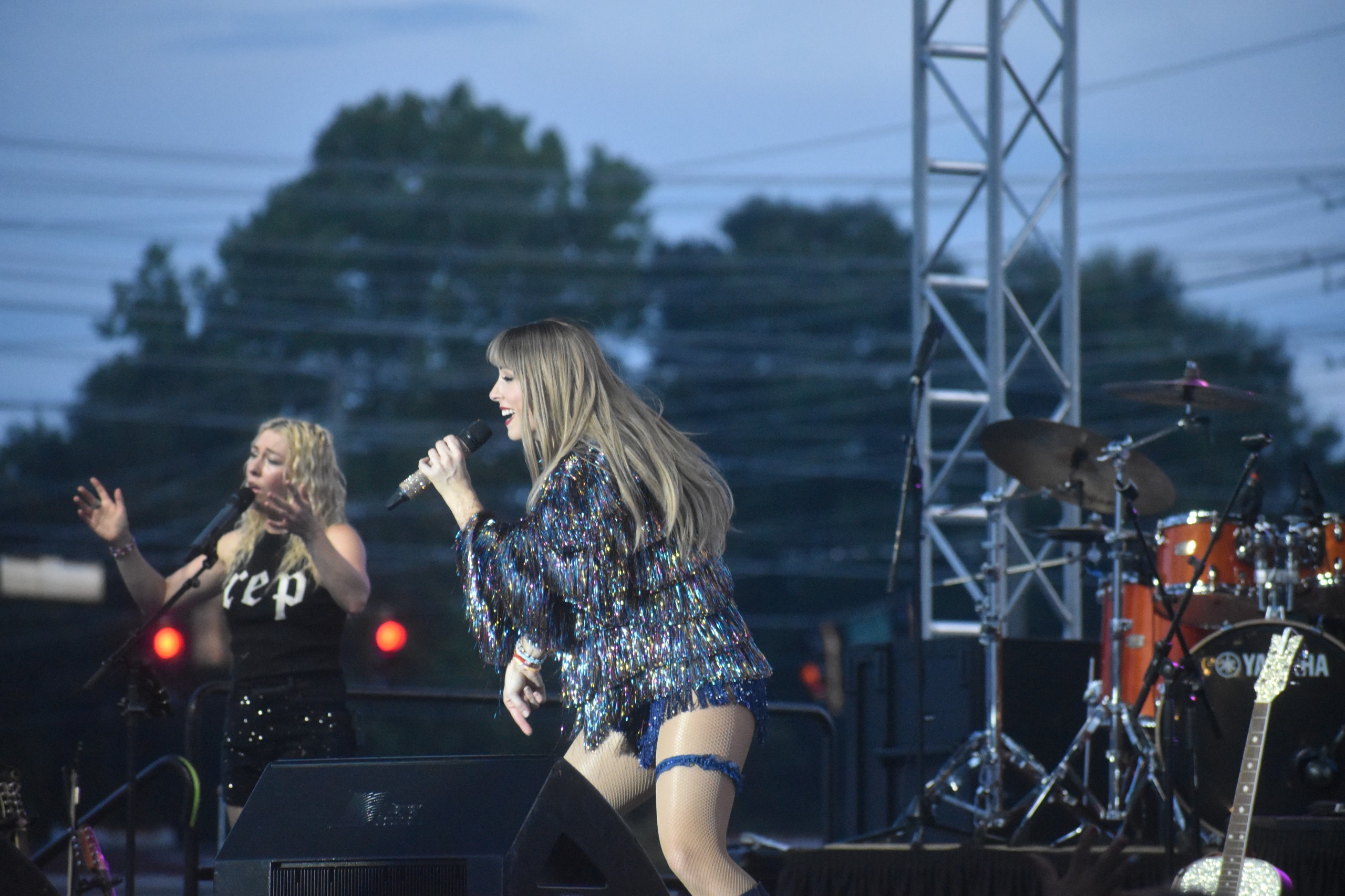 A women performing on stage. She is dressed as famous singer/songwriter Taylor Swift. She is holding a microphone and smiling at the crowd.
