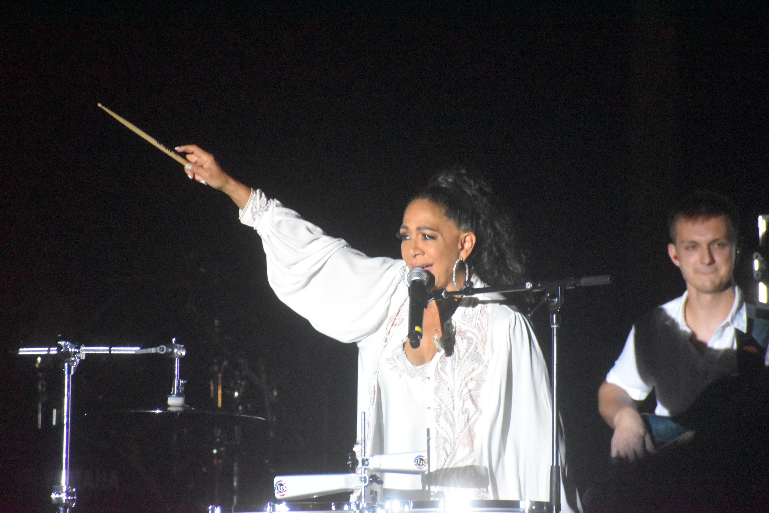 An older women performing. She is wearing an all white outfit. She is holding a drumstick in the air and singing into the microphone.