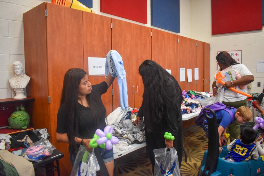 Two women looking at clothes. A woman is also seen in the background. A child is seen speaking to a smaller child who is seated in a cart. 