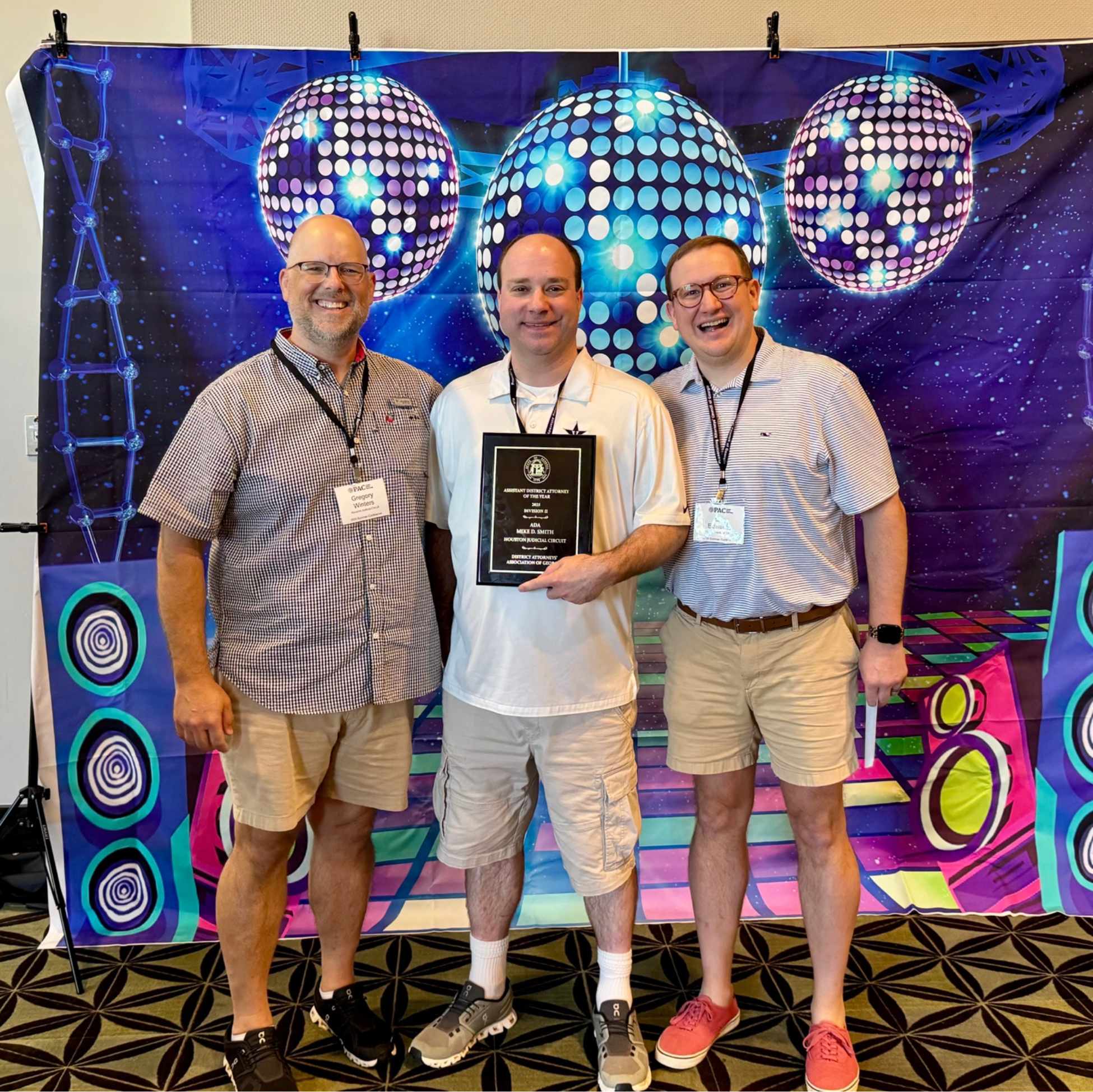 three men wearing shorts standing in front of disco-ball themed backdrop. Man in the middle holding a plaque.