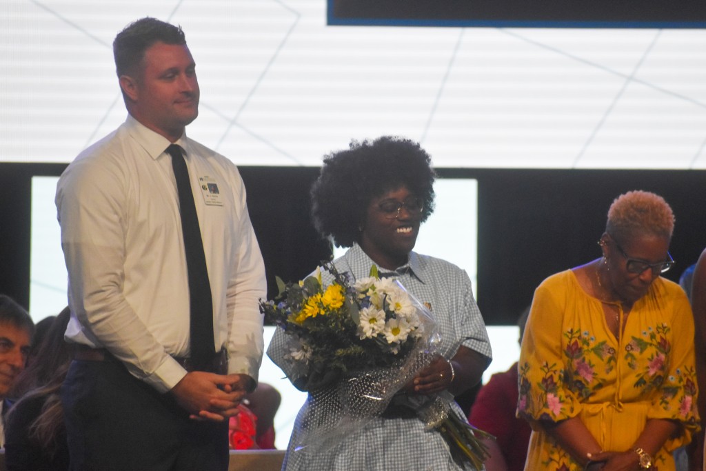A woman smiling and holding flowers. A taller man is standing on her left and a shorter woman is standing on her right.