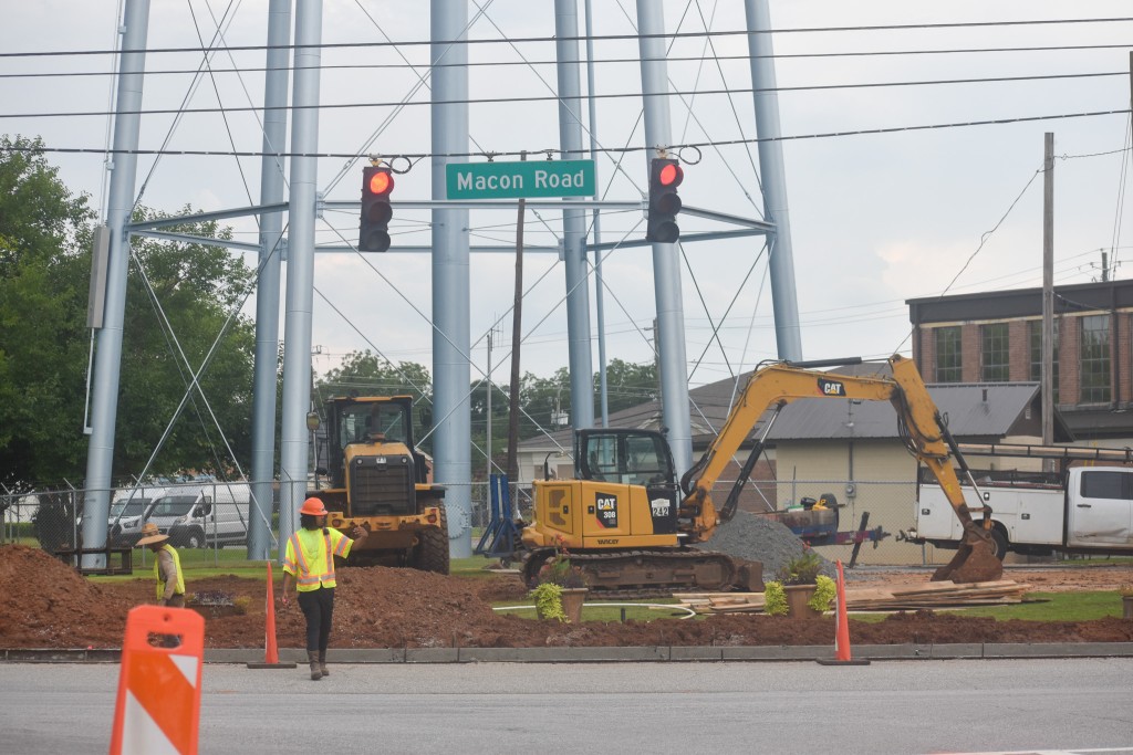 A man in a construction uniform and hard hat on. Above him is a traffic signal and a sign with the words "Macon Road" written.