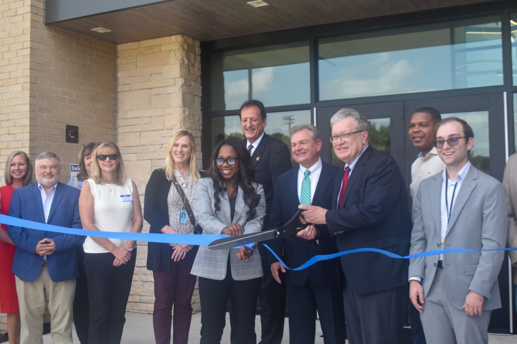 A group of people smiling and standing behind a blue ribbon. The ribbon was just cut by large black scissors. 