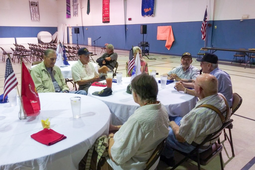 A group of older men sitting at a table. The tables have white cloths over them and the American flag is placed in the middle.