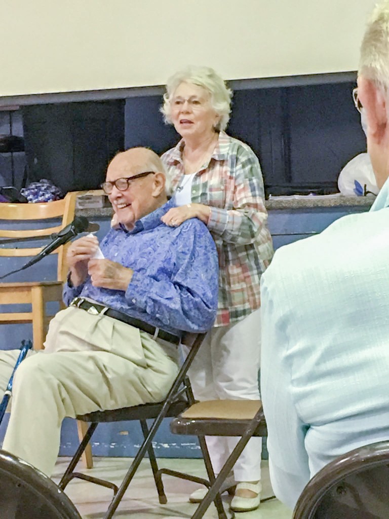 An older man in a chair smiling. A woman is standing behind him speaking to a group of people.