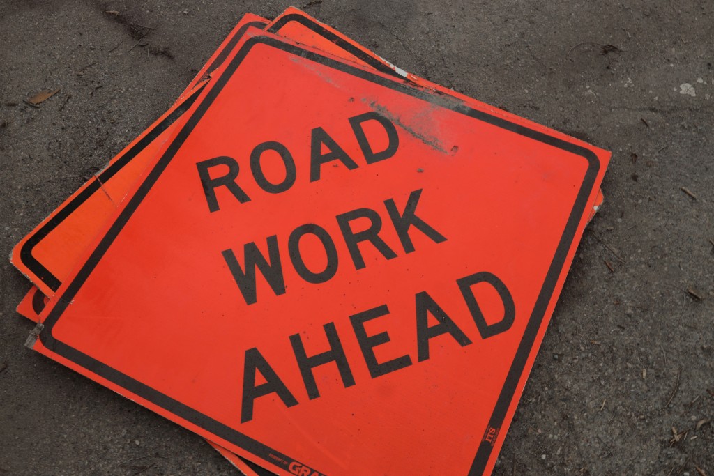 Photo of an orange sign in the shape of a diamond. It says, "road work ahead" written in black letters.