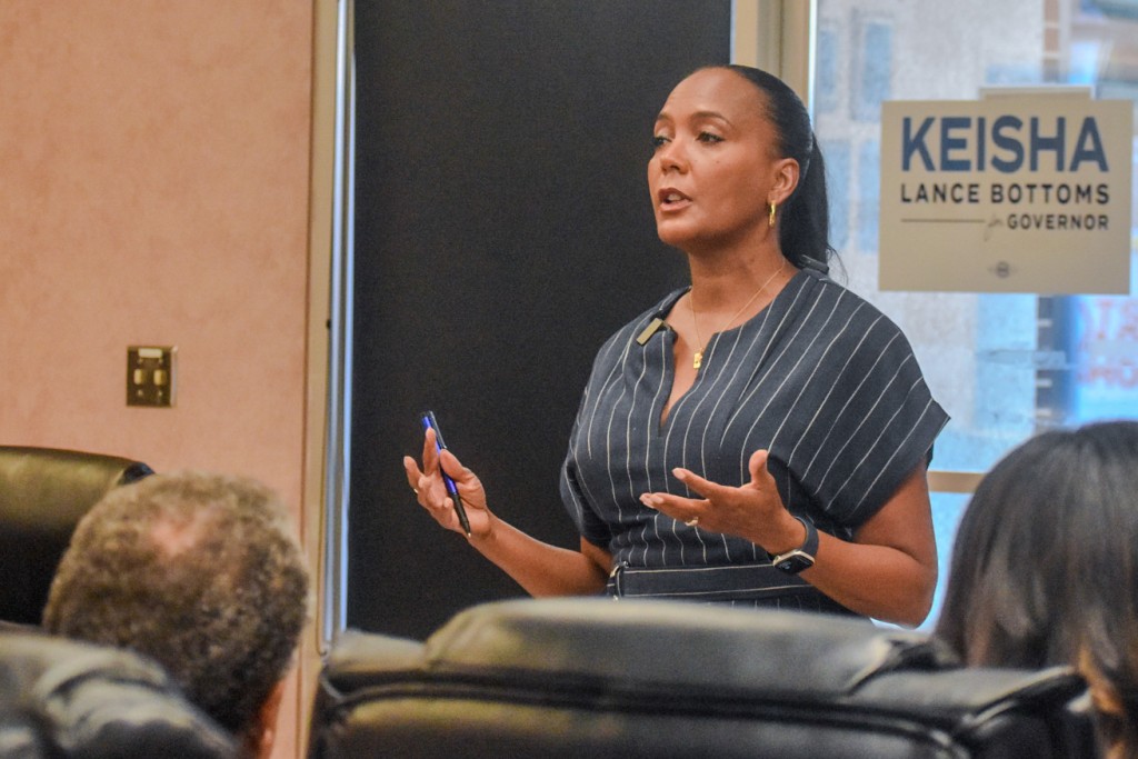 A woman in a dark blue dress speaking to an audience. A campaign sign is seen in the background with "Keisha Lance Bottoms for Governor" written in blue.
