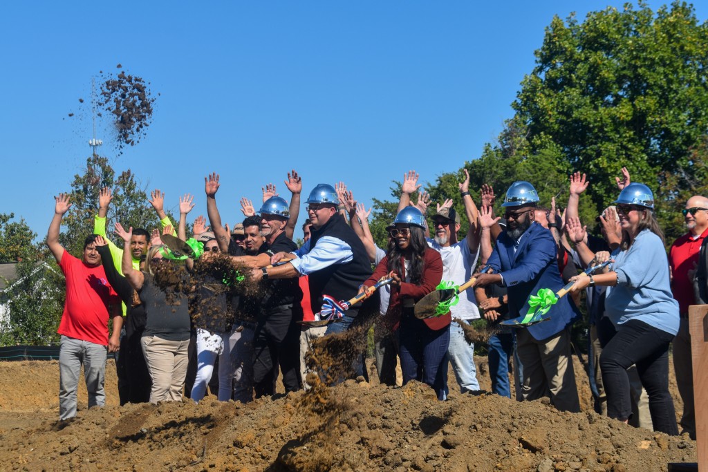 A group of people standing in dirt. They are holding shovels and digging. 