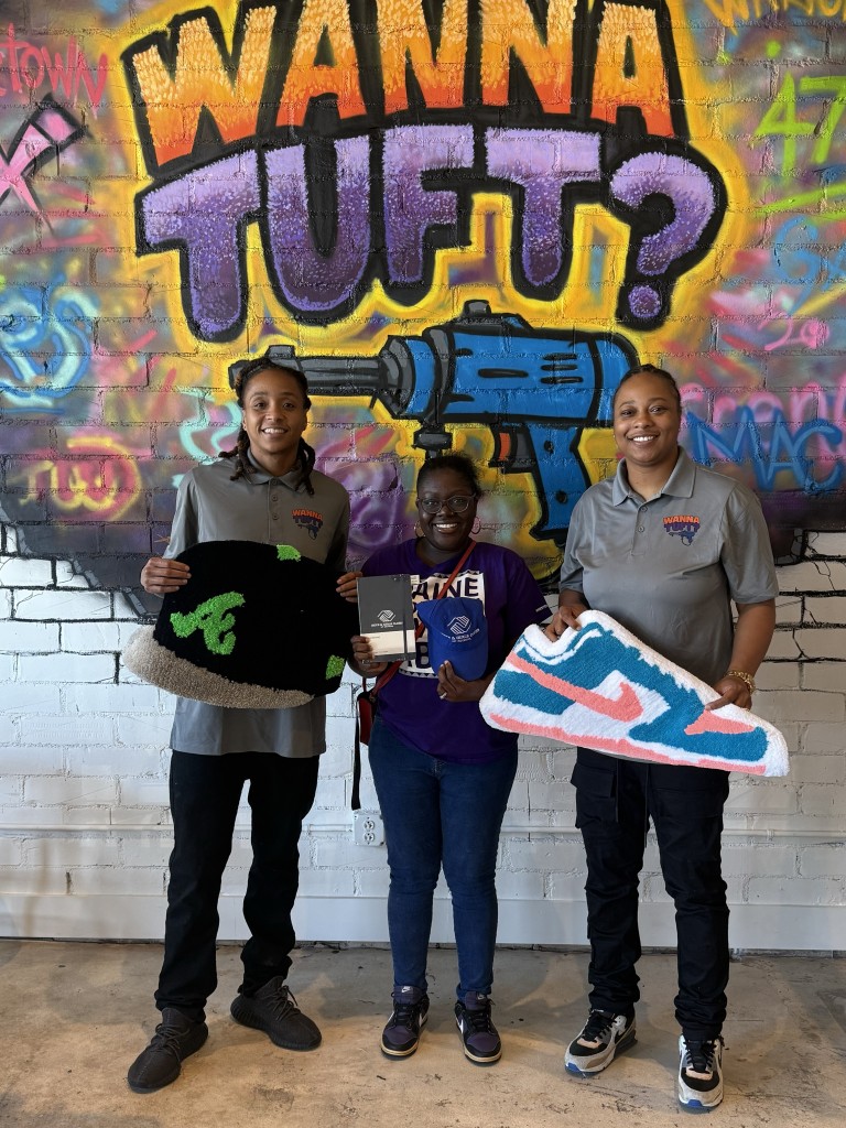 Three women holding rugs. The women on the left is wearing a gray shirt and holding a black rug shaped as a baseball hat. The women on the right is also wearing a gray shirt and is holding a rug shaped as a Nike shoe. A girl is standing in the middle holding Boys and Girls Merchandise.