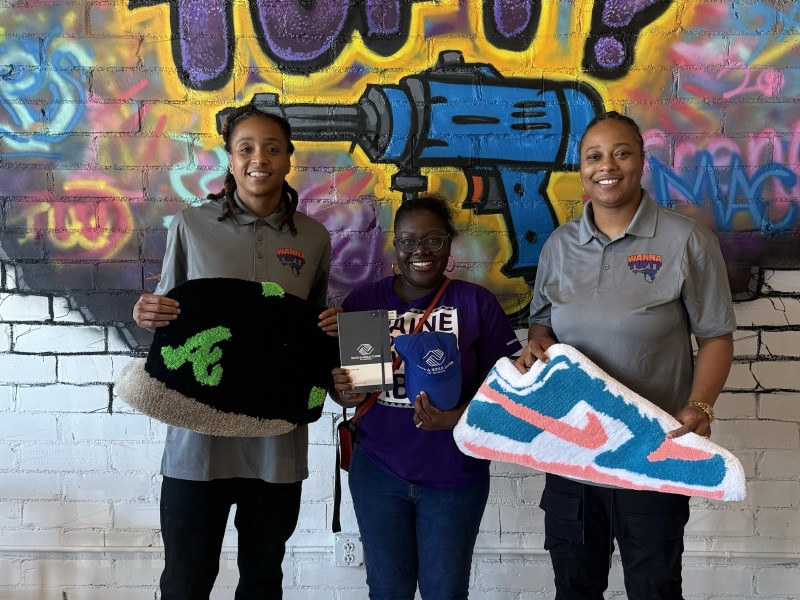 Three women holding rugs. The women on the left is wearing a gray shirt and holding a black rug shaped as a baseball hat. The women on the right is also wearing a gray shirt and is holding a rug shaped as a Nike shoe. A girl is standing in the middle holding Boys and Girls Merchandise.