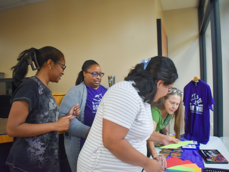 Four women standing over a table which has colored paper and markers.