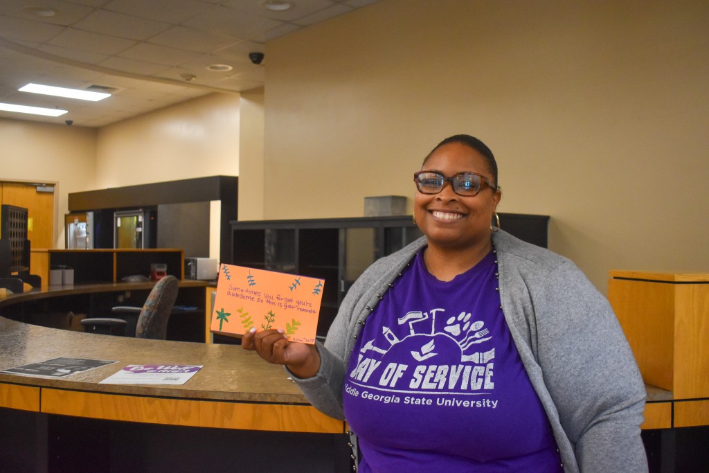 An African American woman in a purple shirt and gray jacket. She is holding an orange card.