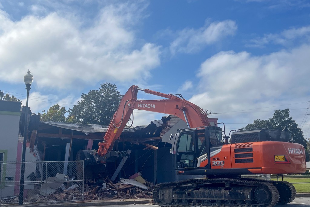 An orange excavator in front of a rumbles of a building. 
