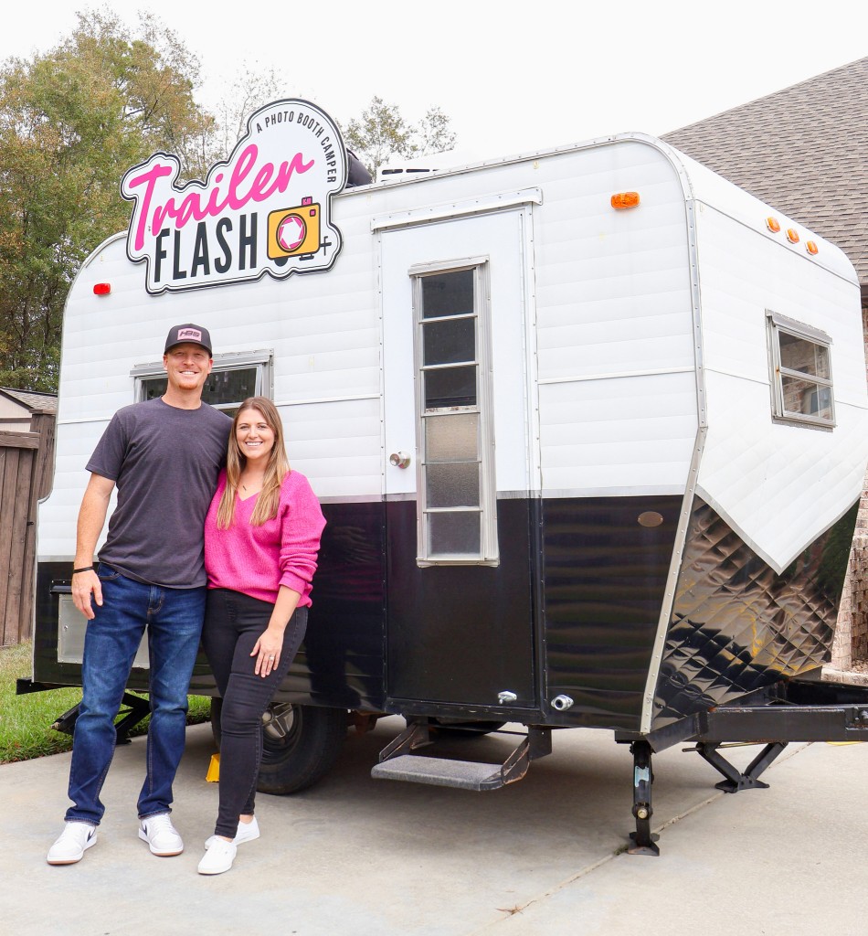 A man and a woman standing in front of a trailer. Both are smiling. The trailer has a sign that says "Trailer Flash" with a yellow camera next to it. 