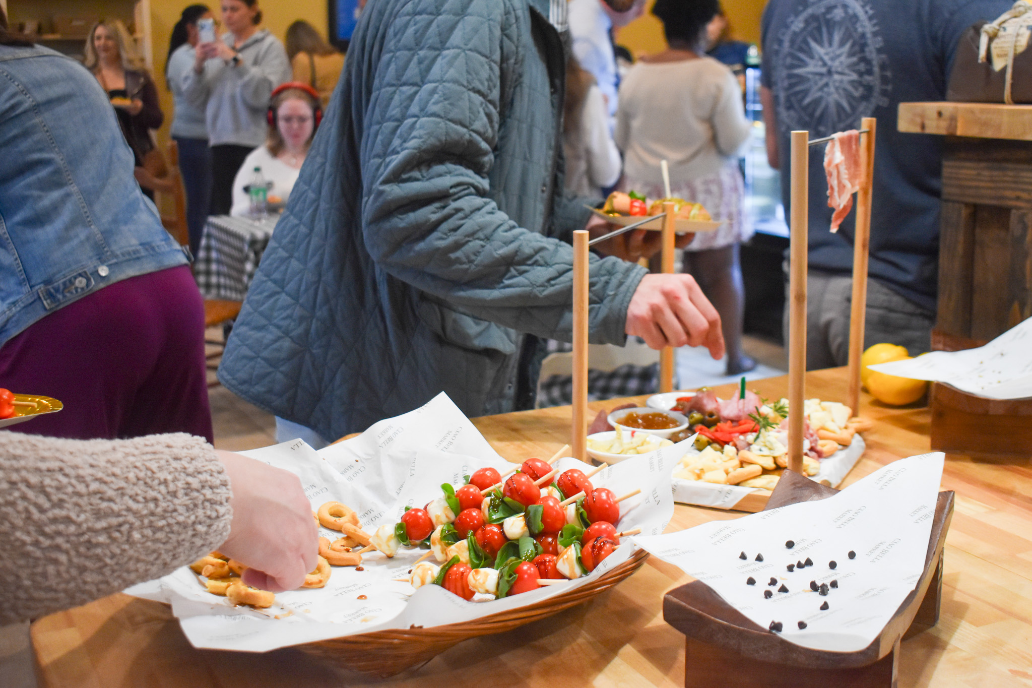A table with food. On one plate is cheese and meets. On the right, there are tomatoes and other vegetables on a stick. There are hands grabbing then food.