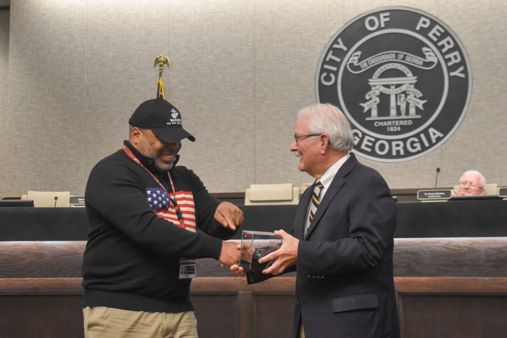 The man on the left is wearing a black hoodie with an American flag on it. He is also wearing a black hate. The man on the right is wear a suit. Both are smiling and holding a box. The man on the left is pointing at the box.