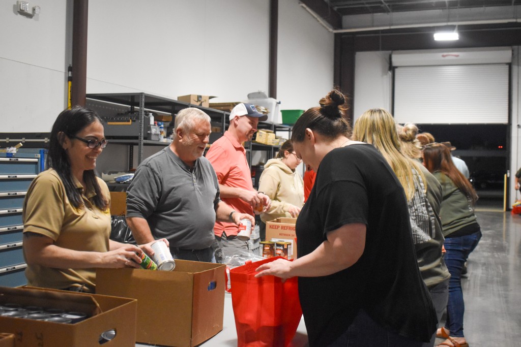 A group of people with a table in the middle. The woman on the left is holding a can. The woman across from her is holding a red bag. The men next to them are smiling.