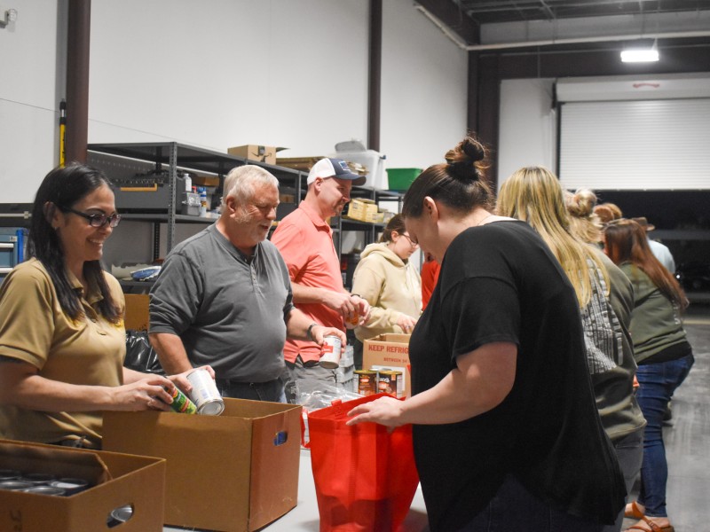 A group of people with a table in the middle. The woman on the left is holding a can. The woman across from her is holding a red bag. The men next to them are smiling.
