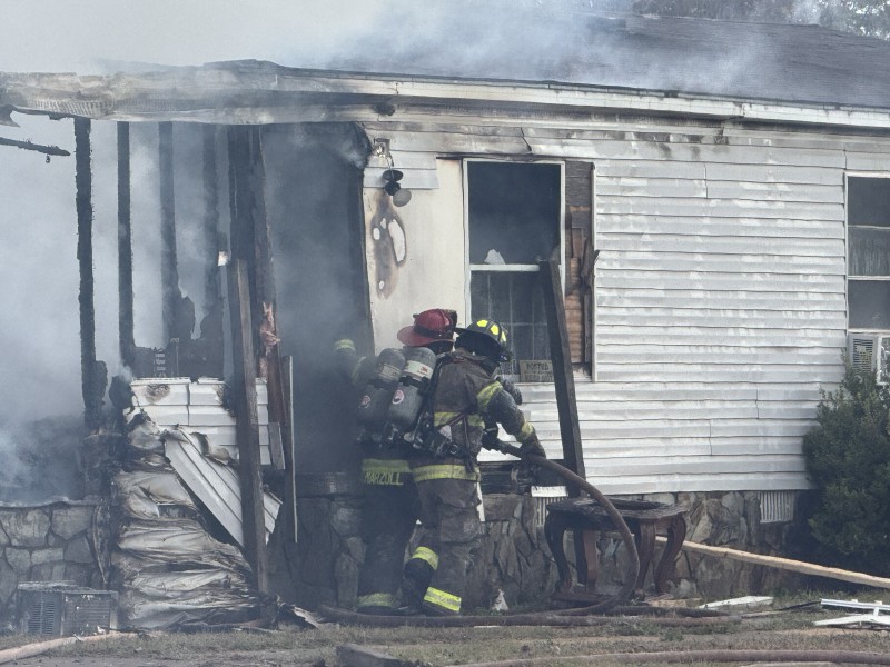 Two people in fire gear. They are standing in front of a house with fire damage. There is still smoke.