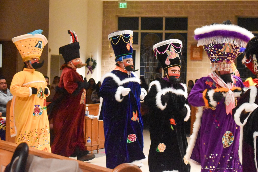 Performers dressed in colorful clothing. Their faces are covered with masks and they wearing large hats. 