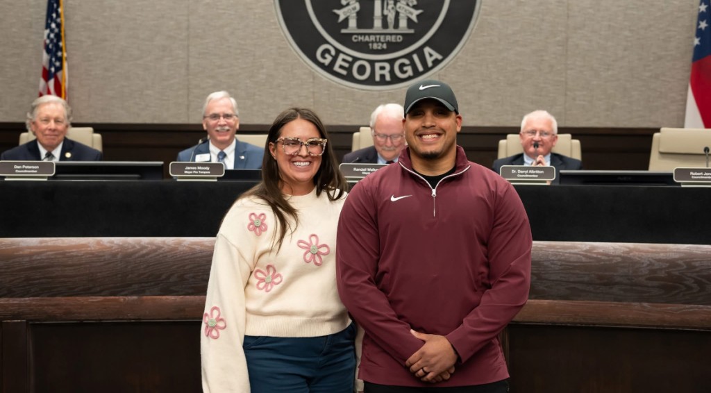 A women in a white sweater and pink flowers standing next a taller man wearing a maroon long sleeve shirt. Both are smiling.