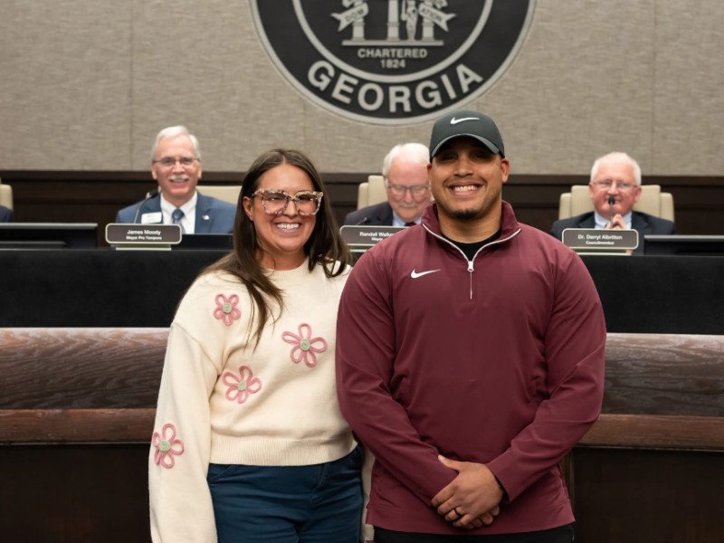 A women in a white sweater and pink flowers standing next a taller man wearing a maroon long sleeve shirt. Both are smiling.