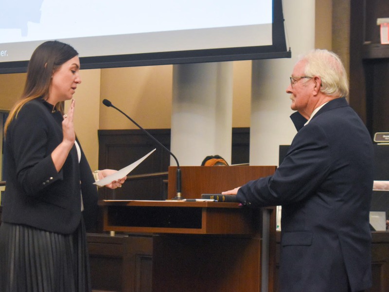 An older man in a black suit raising his right hand and his other hand is on a bible. A woman is standing across from him reading off a paper.