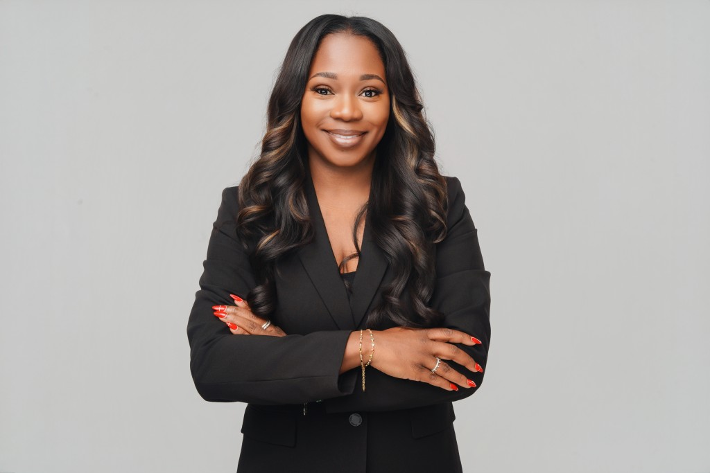 An African American woman smiling. Her arms are crossed and there is a gray background. She is wearing a black suit. 
