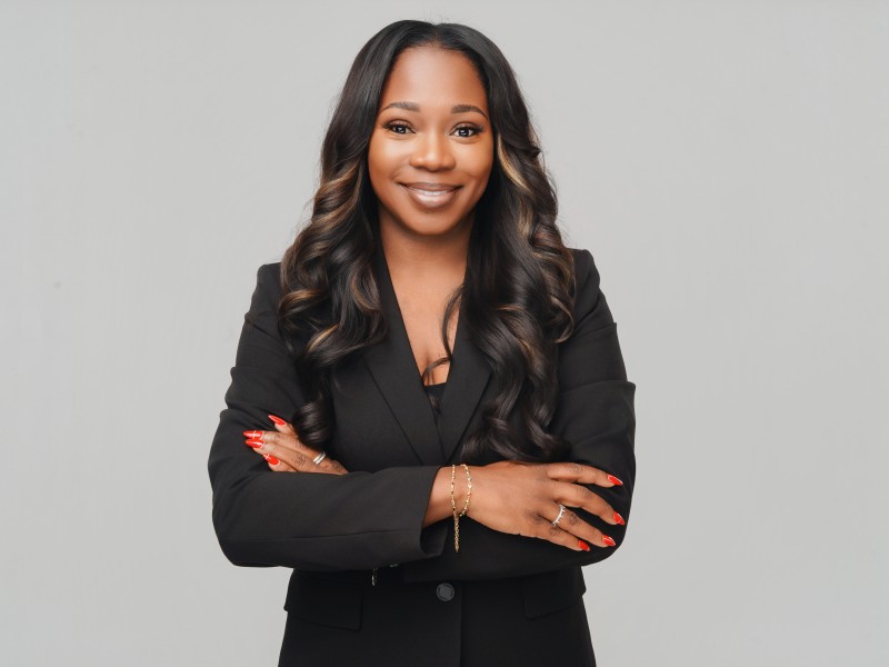 An African American woman smiling. Her arms are crossed and there is a gray background. She is wearing a black suit.