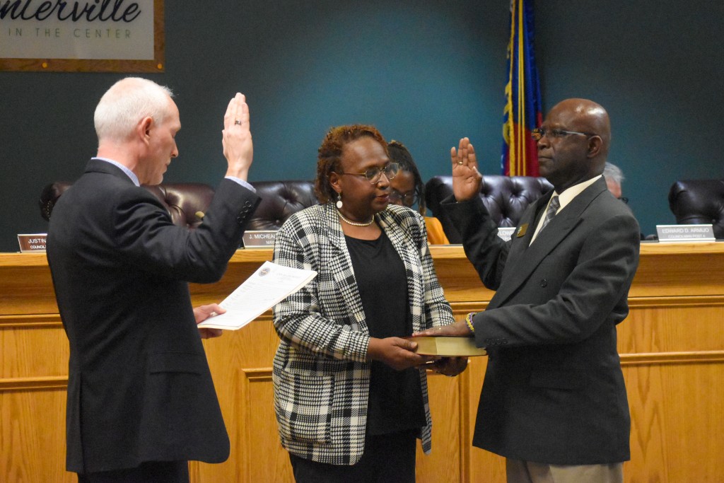 Two men standing across from each other raising their right hand. One of the men has their left hand on the Bible. A woman is standing between them holding the Bible.