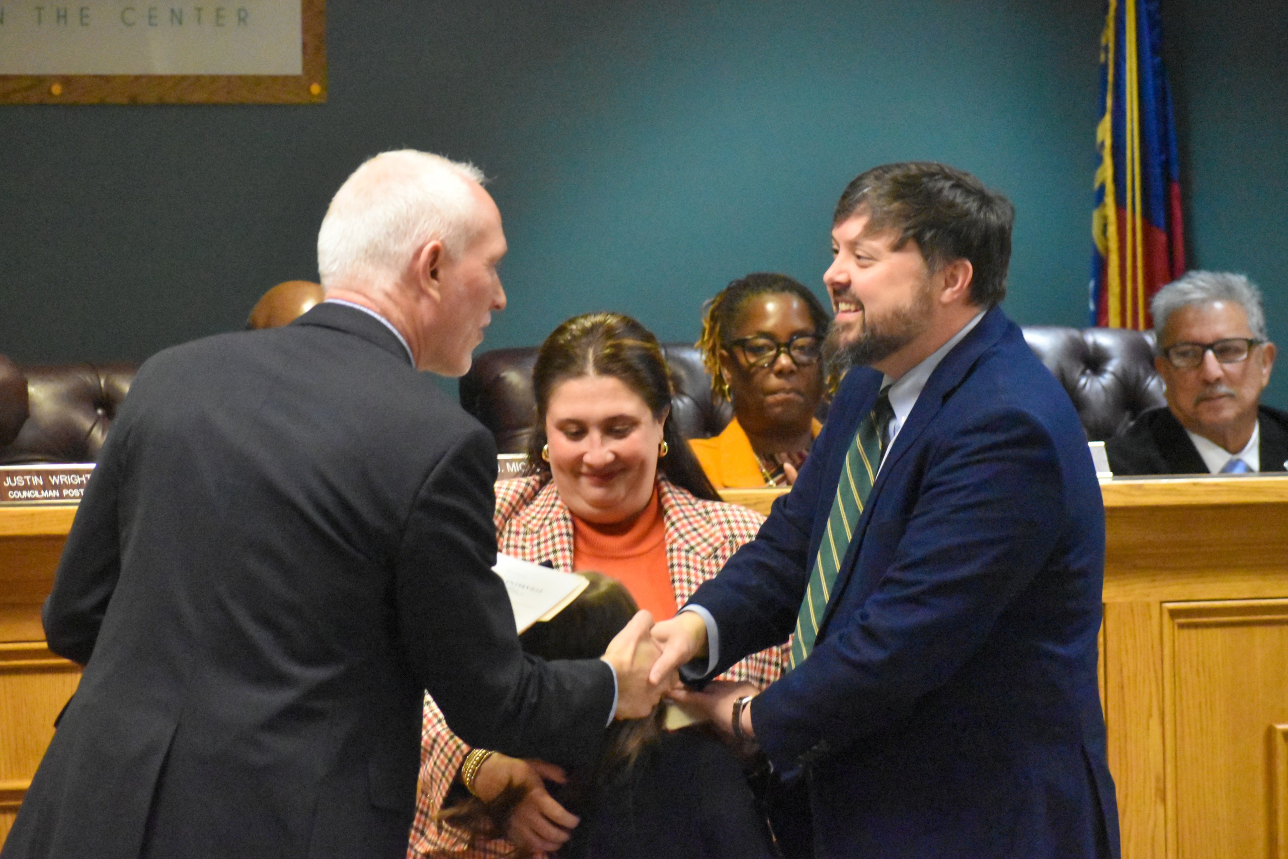 Two men standing across from each other shaking hands and smiling. A woman is standing between them.