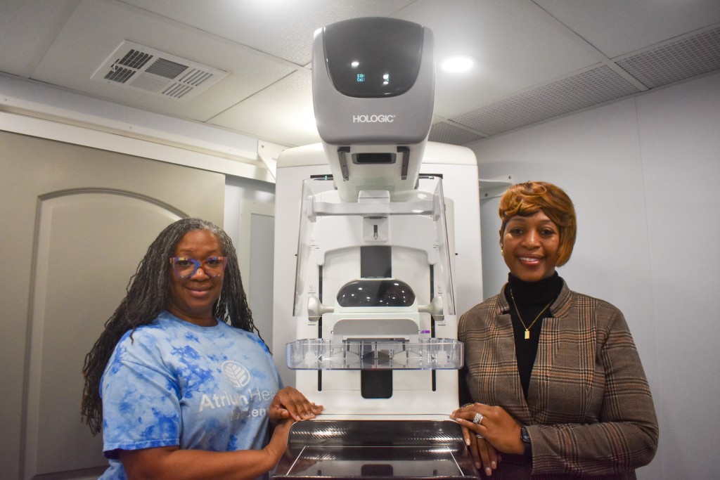 Two women smiling. The women on the left is wearing a blue and white shirt. The woman on the right is wearing a black shirt with a brown coat.
