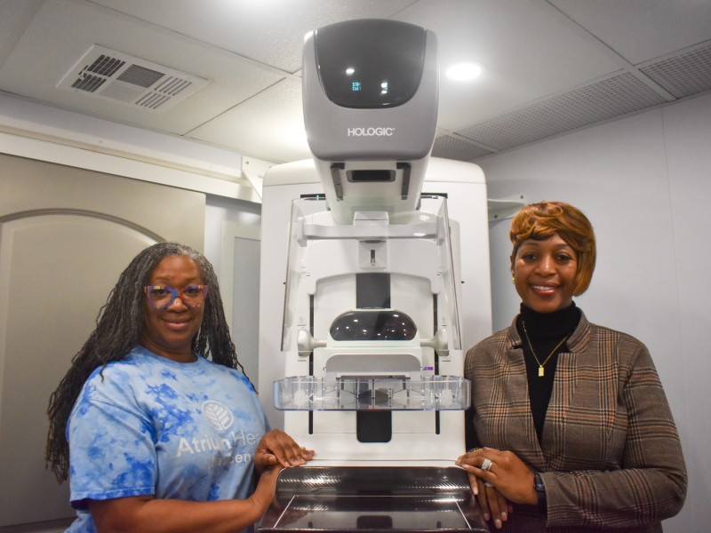 Two women smiling. The women on the left is wearing a blue and white shirt. The woman on the right is wearing a black shirt with a brown coat.