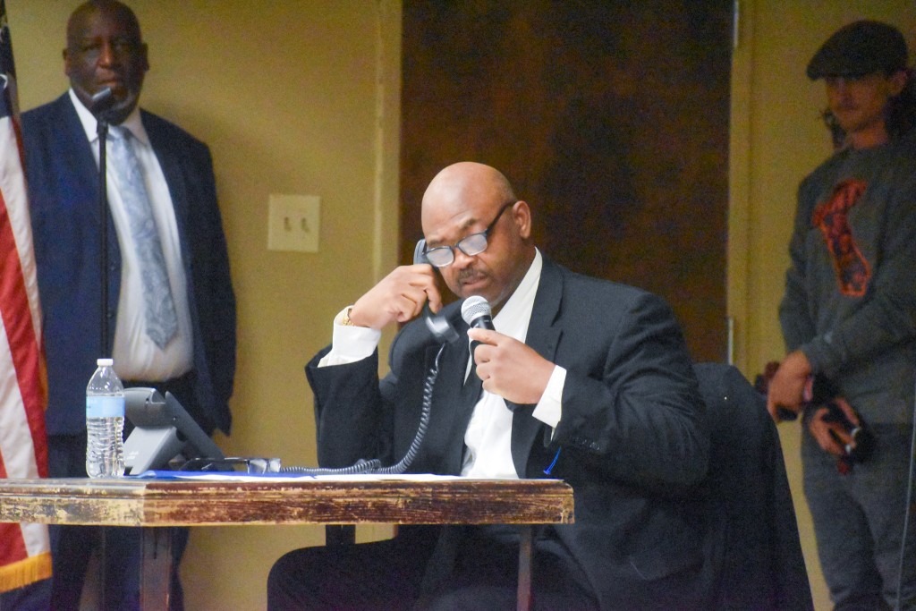 A man in a suit sitting at a table holding a microphone. 