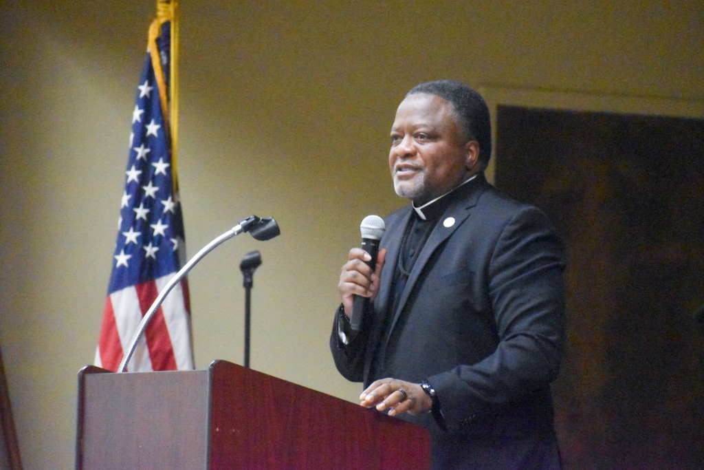 A man in a black suit speaking into a microphone. He is standing behind a podium.
