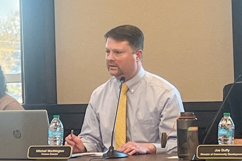 A man in a blue shirt and yellow tie speaking into a microphone. He is sitting behind a desk.