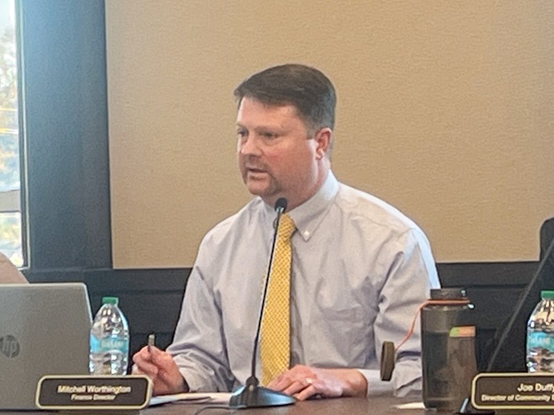 A man in a blue shirt and yellow tie speaking into a microphone. He is sitting behind a desk.